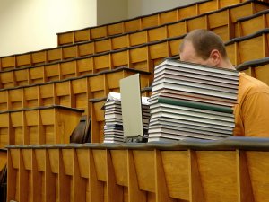 Student in lecture hall with face hidden behind books and laptop