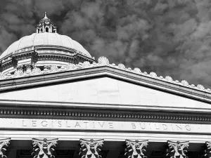 Black and white exterior of Legislative chambers of the Washington State Capitol in Olympia with inscription, pillars, and dome with a cloudy sky in the background.