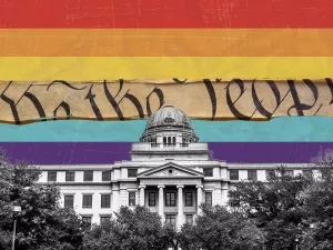 Pride Flag with one stripe removed revealing the words "We the people" from the Constitution with the Texas A&M Academic Building in the foreground.