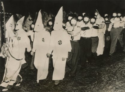 Ku Klux Klan men in full white masks and gowns lead new members wearing small face masks
