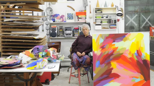 Artist Samia Halaby sitting on a chair in her studio next to a large abstract painting